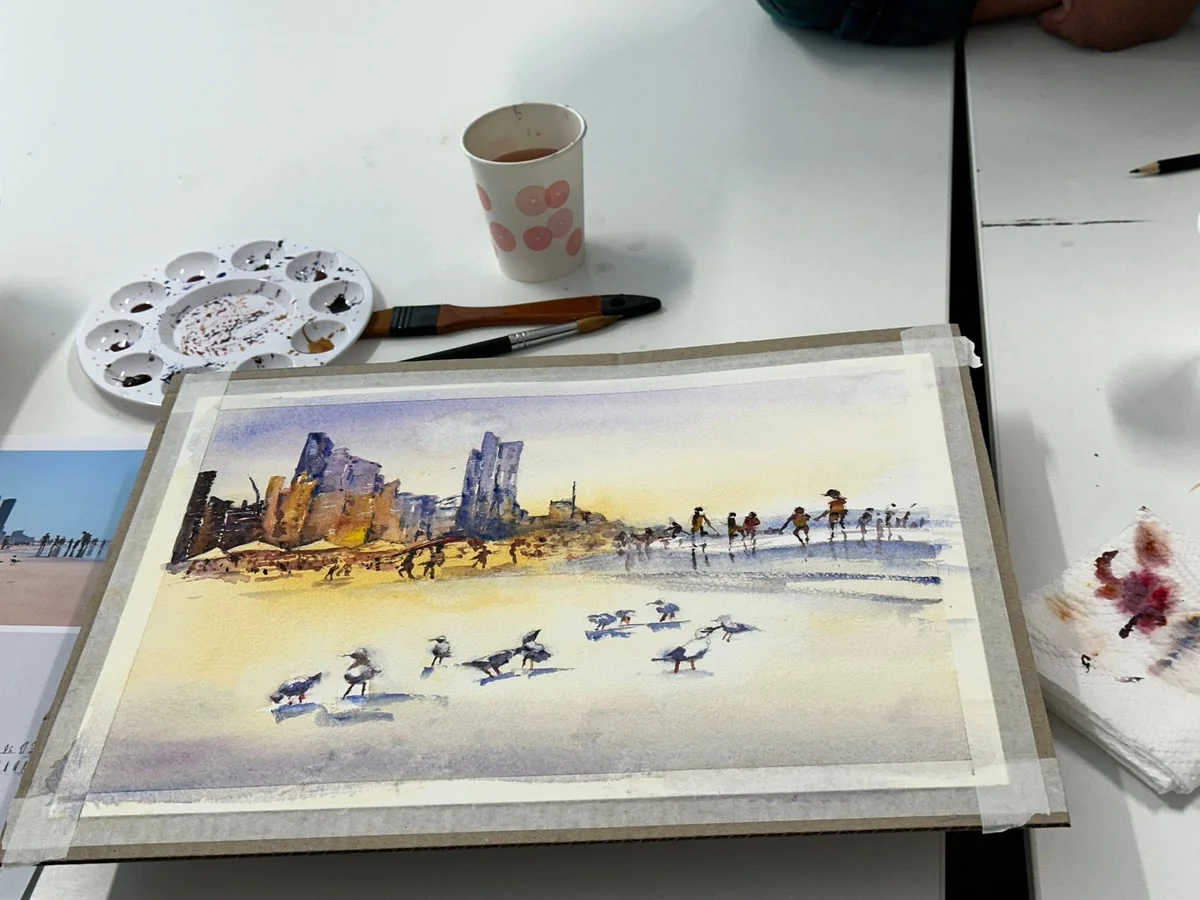 Watercolour of a beach and city skyline with seagulls, taped to a board beside a palette, brushes and reference photograph