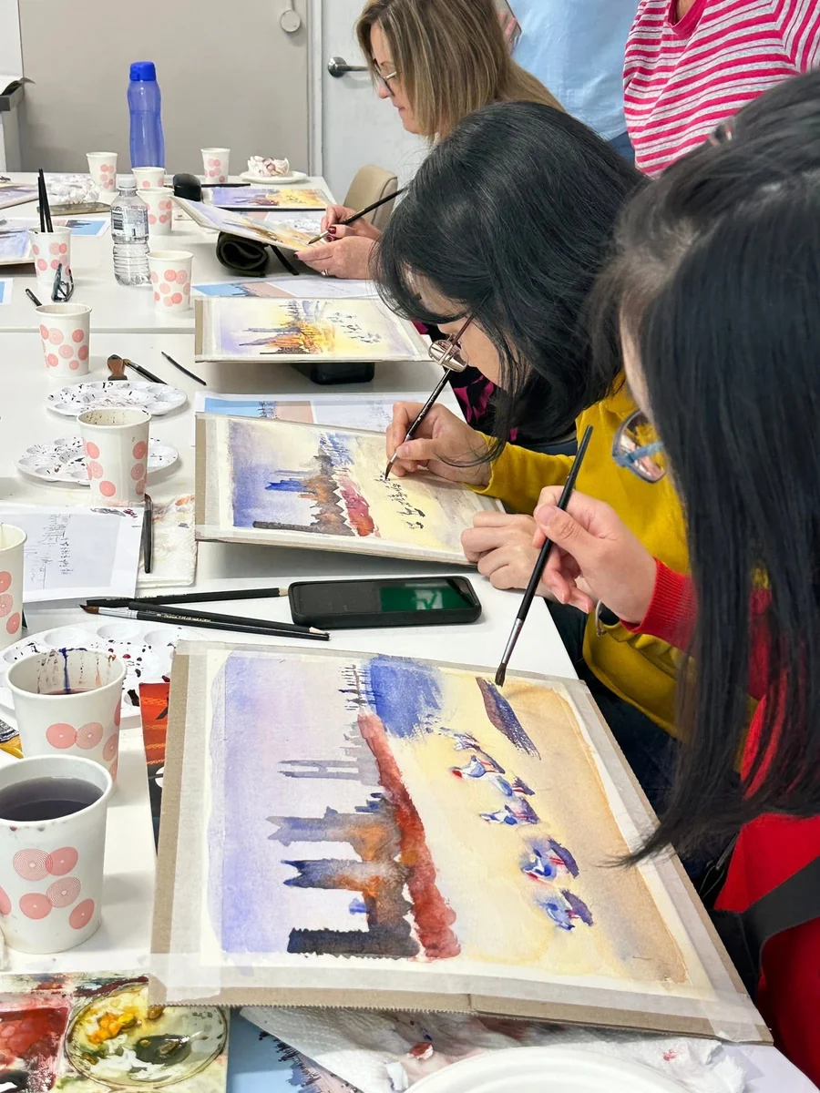 Participants seated along the table painting watercolour skylines, brushes in hand, palettes and reference prints beside them