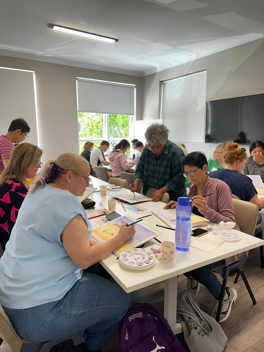 Participants working at joined tables with palettes, brushes and paper, an assisting participant leaning over to look at a painting